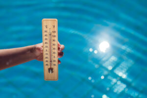 Thermometer displaying temperature in Celsius and Fahrenheit held above a swimming pool with sunlight reflecting on the water.